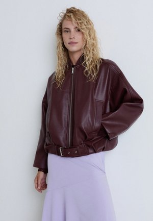 Blonde Frau trägt eine bordeauxrote Lederjacke mit Reißverschluss und Gürtel-Detail sowie einen hellvioletten Rock, steht vor einem schlichten weißen Hintergrund.