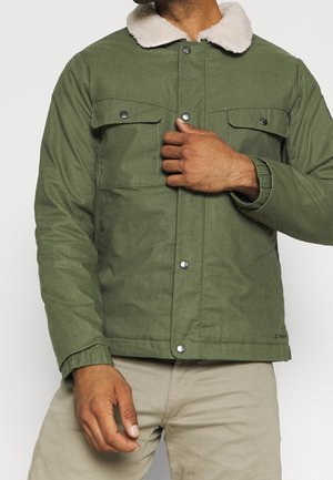 Mann trägt eine olivgrüne Jacke mit weißem Fleecefutter am Kragen und Druckknöpfen, kombiniert mit beigen Hosen, steht vor einem schlichten Hintergrund.