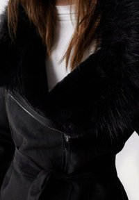 Mujer con un abrigo negro con un gran cuello de piel y cremallera, sobre una blusa blanca, con el cabello castaño suelto visible a la altura de los hombros.