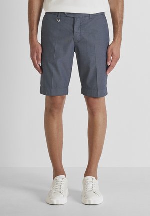 Hombre con pantalones cortos grises a medida con dobladillos remangados y zapatillas blancas, de pie contra un fondo gris claro liso.