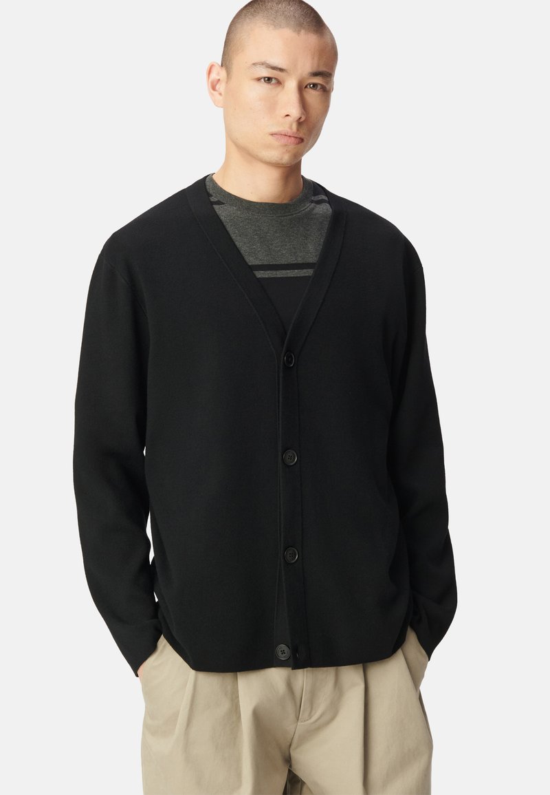 Cárdigan negro con escote en V, botones en la parte delantera y mangas largas, combinado con una camisa gris. El tejido parece suave y de punto. Pantalones beige claro visibles.