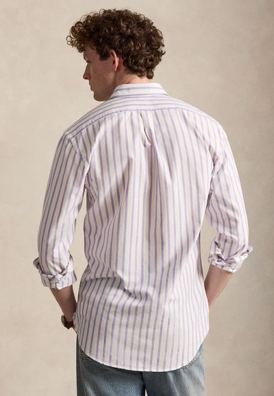 Mann mit lockigem Haar, der ein weißes Hemd mit vertikalen violetten Streifen trägt, die Ärmel hochgekrempelt hat und blaue Jeans anhat, mit dem Rücken zur Kamera, die Hände in den Taschen.