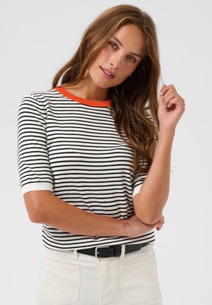 Femme aux longs cheveux bruns portant une chemise rayée noire et blanche avec un col orange et un pantalon clair avec une ceinture noire.