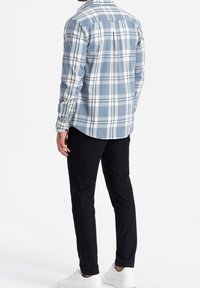 Camisa de cuadros azul y blanca con mangas largas y puños abotonados, combinada con pantalones negros y zapatillas blancas. La camisa tiene un dobladillo curvo.