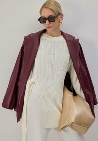 Veste blazer en similicuir bordeaux portée sur un pull en maille torsadée blanc, associée à un pantalon crème et une pochette en satin beige. Lunettes de soleil noires carrées.