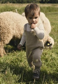 Beige gestrickte Latzhose mit strukturiertem Muster, Knopfdetails und langärmeligem weißen Hemd, vor einem grünen Feld mit Schafen im Hintergrund.