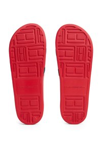 Semelles de chaussures en caoutchouc rouge avec des motifs texturés et le logo "TOMMY HILFIGER" en relief. Bords arrondis et designs géométriques distincts tout au long.