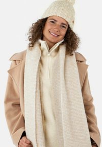 Manteau en mélange de laine beige avec de larges revers, pull en maille côtelée crème et écharpe en crème texturée. Un bonnet en tricot avec un pompon complète la tenue.