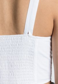 Close-up of a person’s back wearing a white smocked top with wide shoulder straps against a plain background.