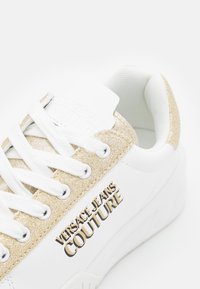 Baskets blanches avec des détails en paillettes dorées, des lacets blancs et le logo "Versace Jeans Couture" sur le côté en lettres noires et dorées.