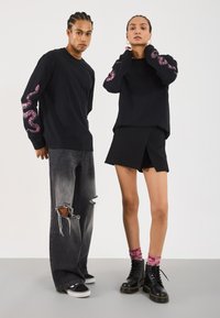 Chemise noire à manches longues avec imprimé serpent rose sur les manches, associée à un jean gris déchiré et une jupe noire. Présente des bottes noires à lacets.