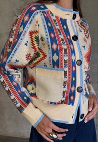 Γυναίκα που φοράει πολύχρωμη πλεκτή ζακέτα με μεγάλα κουμπιά, μπεζ τσέπη και μπλε πινελιέ ς, που στέκεται μπροστά σε γκρίζο τοίχο.