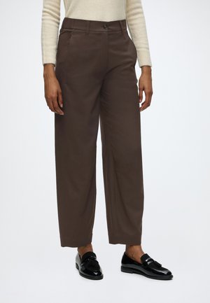 Woman wearing dark brown wide-leg trousers, cream long-sleeve top, and black shiny tassel loafers standing on white background.