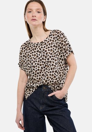 Femme portant une chemise beige à manches courtes imprimé léopard, rentrée dans un jean bleu foncé, debout avec une main dans la poche.