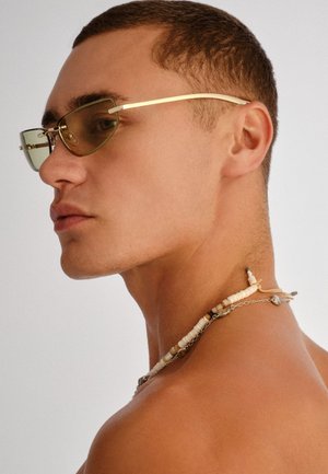 Jonge man met kort haar, die geel getinte zonnebril en een kralenketting met schelpen en metalen hangers draagt, kijkt naar links.