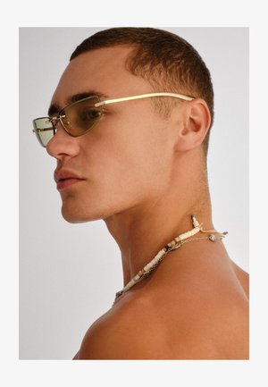 Mladić s kratkom kosom, koji nosi sunčane naočale žućkaste nijanse i ogrlicu od perli s školjkama i metalnim privjescima, gleda ulijevo.