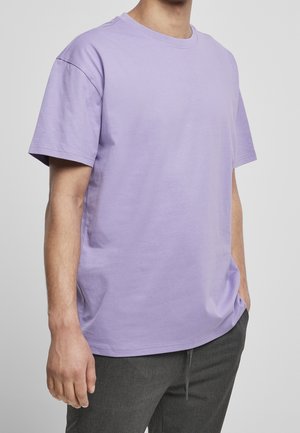 Mand iført en ensfarvet lavendel kortærmet T-shirt og mørkegrå bukser, stående med den ene hånd i lommen mod en lysegrå baggrund.