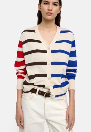 Kobieta w beżowym kardiganie z asymetrycznymi czerwonymi, brązowymi i niebieskimi poziomymi paskami, w połączeniu z białymi spodniami i brązowym paskiem.