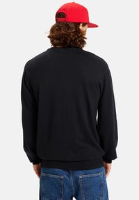 Black knit pullover with long sleeves, featuring a round neckline and ribbed hem, paired with blue denim jeans and a red cap.