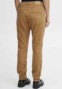 Pantalons en mélange de coton marron avec une coupe fuselée, dotés de deux poches arrière et d'une étiquette à la taille. Texture lisse et design décontracté.
