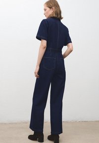Žena stojaca bokom, oblečená v tmavomodrom denimovom overal s krátkymi rukávmi, širokými nohavicami a čiernymi členkovými čižmami na podpätku.
