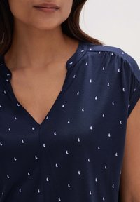 Laivastonsininen paita, jossa on V-kaula-aukko ja hihattomat hihat, eläviä valkoisia perhoskuvioita ympäri. Pehmeä, sileä kangas, jossa on hienovarainen kiilto.