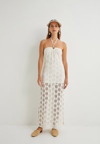 Weißes gehäkeltes Maxikleid mit Halterneck und Schlitzdetail. Durchscheinender Stoff mit Blättermustern. Modell trägt geflochtene Slippers.