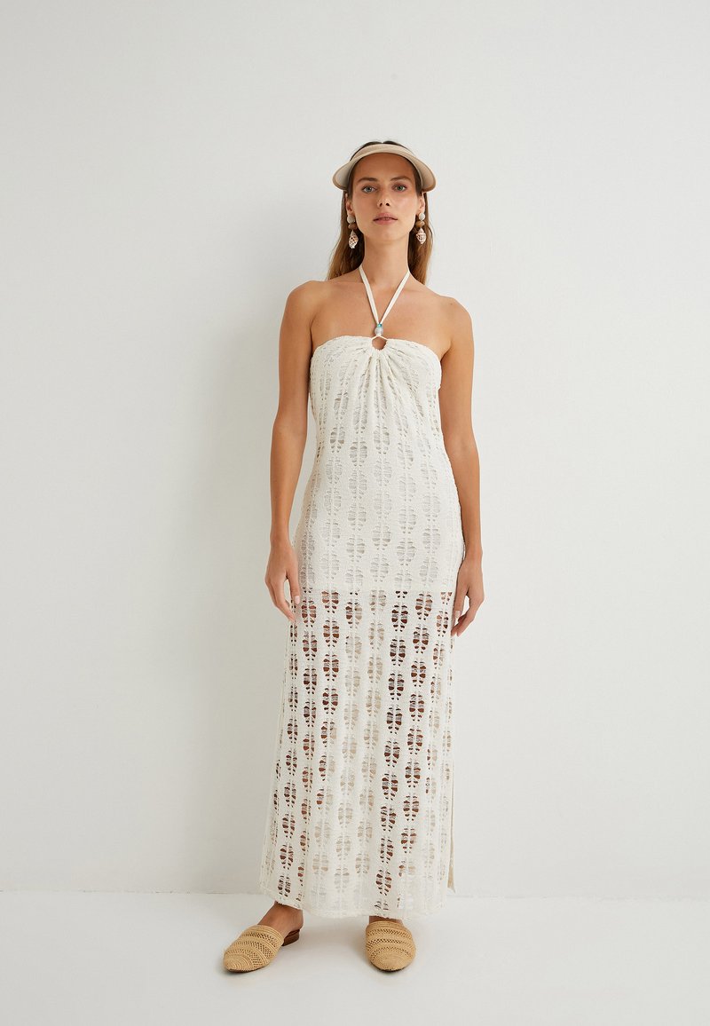 Weißes gehäkeltes Maxikleid mit Halterneck und Schlitzdetail. Durchscheinender Stoff mit Blättermustern. Modell trägt geflochtene Slippers.