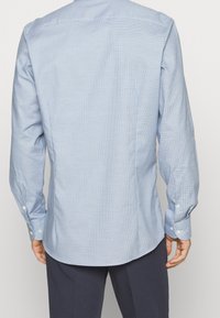 Uomo che indossa una camicia da uomo a maniche lunghe di un blu chiaro con un sottile motivo a griglia e pantaloni scuri, visto da dietro.