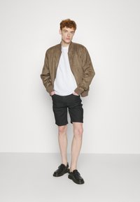 Bomber jacket de ante marrón con puños acanalados, camiseta blanca, pantalones cortos oscuros y zapatos negros sin cordones. El modelo está de pie con las manos en los bolsillos.