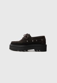 Scarpe scure in suede marrone con una robusta suola nera. Presentano lacci, occhielli metallici e un design della punta cucito. Vista laterale.