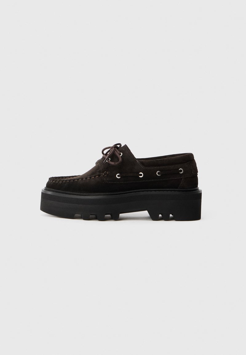 Scarpe scure in suede marrone con una robusta suola nera. Presentano lacci, occhielli metallici e un design della punta cucito. Vista laterale.