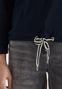 Haut bleu marine côtelé avec cordon à rayures noué à la taille, associé à un jean gris délavé et une main détendue reposant le long du corps.