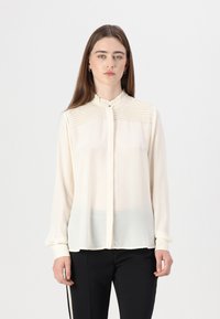 Cream blouse with a ruffled collar, textured detailing on shoulders, and button closure. Long sleeves and sheer fabric design.
