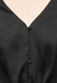 Μαύρο ύφασμα με V-λαιμόκοψη και δύο ορατά μαύρα κουμπιά κατά μήκος του κέντρου μπροστά πάνω σε φωτεινό δέρμα.