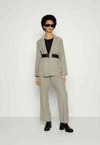 Fato cinza ajustado com um blazer estruturado e calças de perna larga, combinado com um cinto preto, uma blusa de manga longa e botas pretas robustas.