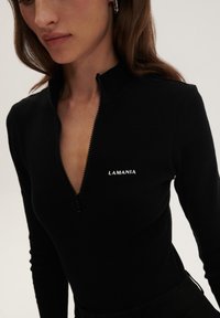 Juodas ilgas rankovėmis marškinėliai su ketvirčio užtrauktuku ir baltu logotipu "LAMANIA". Su aukšta apykakle ir prigludusia forma. Lygi tekstūra.