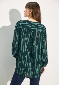 Camicia verde a motivi con maniche lunghe, caratterizzata da un taglio morbido e un tessuto testurizzato che mostra linee e forme verticali.