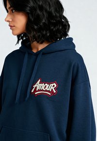 Felpa blu navy realizzata in tessuto morbido, con un logo ricamato "Amour" in rosso e bianco in evidenza. Include una tasca frontale e cappuccio con cordini.