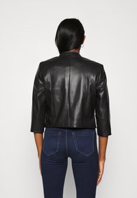 Veste en cuir noir cropped avec des manches 3/4, des coutures structurées et une texture lisse. Portée sur un jean en denim foncé.