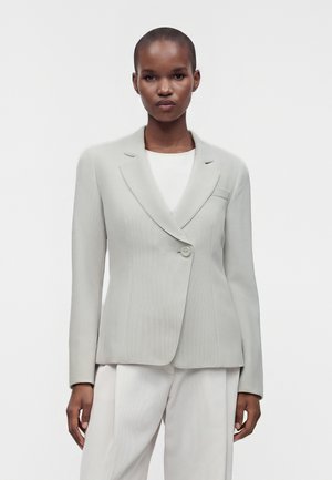 Femme debout devant un fond uni, portant un blazer gris clair ajusté sur un haut blanc et un pantalon assorti, face à l'avant.