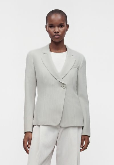 Femme debout devant un fond uni, portant un blazer gris clair ajusté sur un haut blanc et un pantalon assorti, face à l'avant.