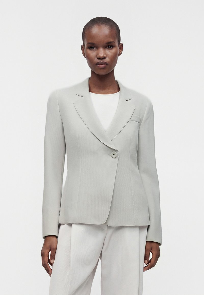 Femme debout devant un fond uni, portant un blazer gris clair ajusté sur un haut blanc et un pantalon assorti, face à l'avant.