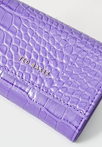 Portafoglio in pelle verniciata viola con texture a coccodrillo. Logo "TED BAKER" in oro impresso sul fronte, caratterizzato da un design elegante e compatto.
