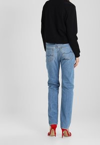 Kobieta ubrana w jasnoniebieskie dżinsy, czarną bluzkę z długim rękawem i czerwone sandały na wysokim obcasie, stoi tyłem do aparatu.