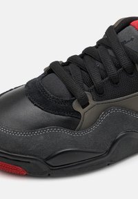 Svart og grå sneaker med en kombinasjon av skinn og semsket skinn, med svarte snører og en rød aksent på sålen.