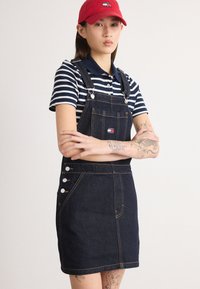 Džinsu kopējais kleita ar regulējamiem siksnām, tumša veidā, priekšējais kabata, sānu pogu akcenti. Apvienots ar tumši zilu un baltu svītrainu polo. Sarkans cepure.