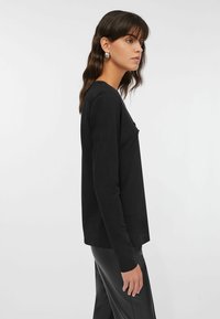 Top nero a maniche lunghe realizzato in tessuto morbido, con scollo rotondo e vestibilità rilassata, indossato con pantaloni neri lucidi.