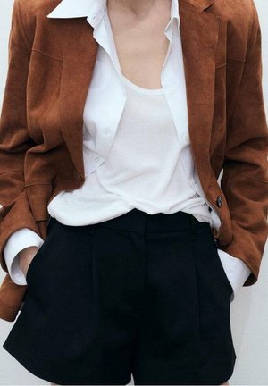 Persoon draagt een bruine blazer over een wit overhemd en een tanktop, ingestopt in zwarte, hooggetailleerde shorts met handen in de zakken.