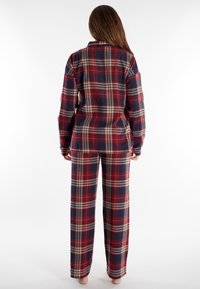 Geruite flanellen pyjama set in rood, marineblauw en crème. Heeft een knoopsluiting bovenstuk met een kraag en lange mouwen, en bijpassende wijde broek.